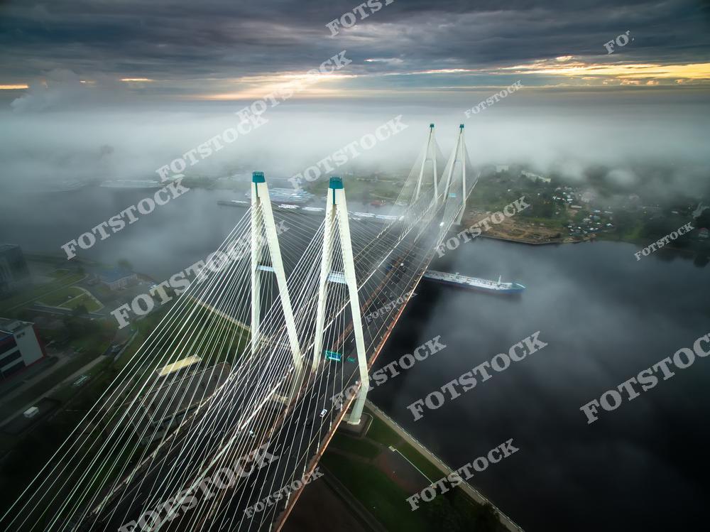 На фотографии изображен Большой Обуховский (Вантовый) мост, соединяющий берега Невы. Густой туман создает мистический эффект, скрывая часть моста и окрестности под белым покрывалом. Утренние лучи света пробиваются сквозь облака, добавляя драматичности в кадр. Вантовый мост, с его изящными опорами и тросами, выглядит монументально и впечатляюще даже в условиях ограниченной видимости.
